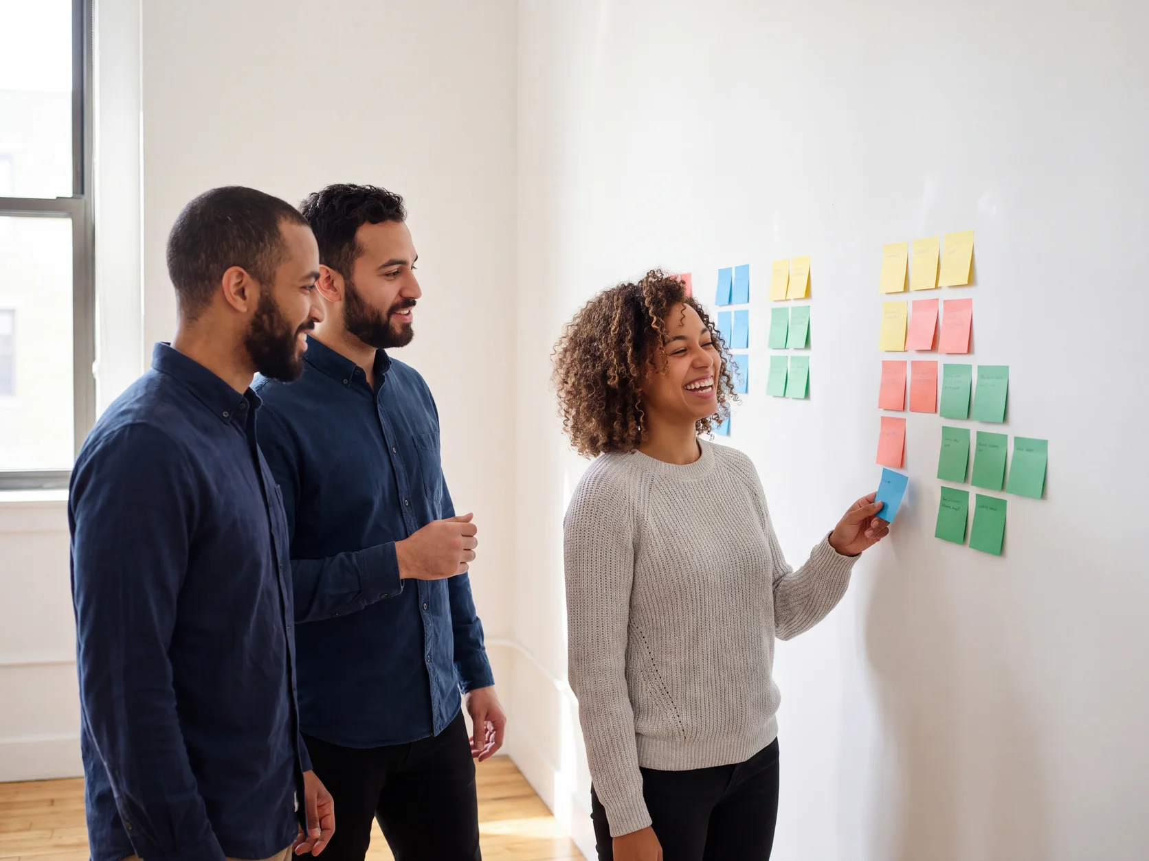 Trois designers regroupent des fiches colorées par thème sur un mur blanc, dans un studio lumineux. Scène générée par IA.